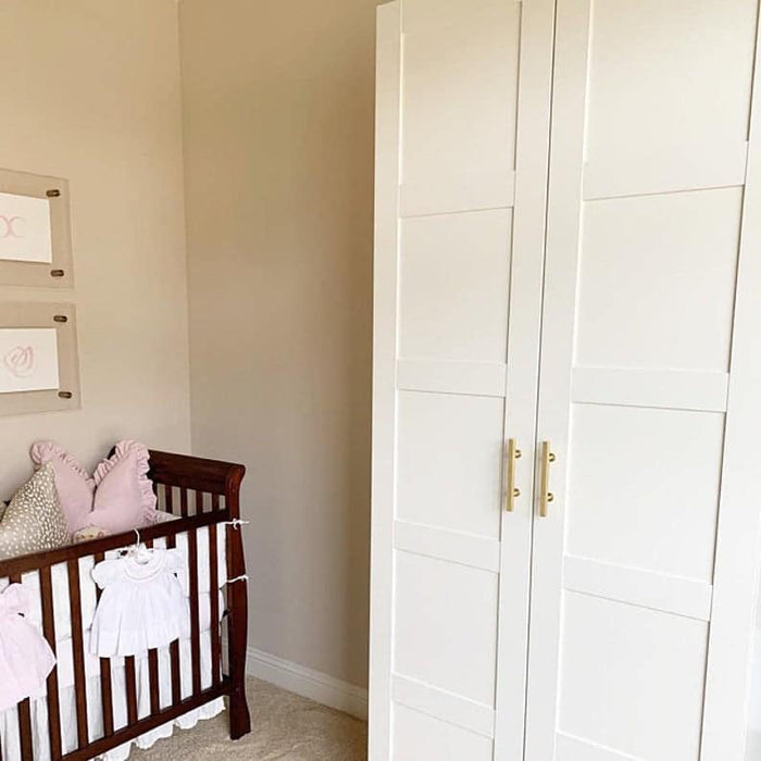 A nursery with a dark wooden crib, pink and white bedding, two wall art pieces, and a large white wardrobe featuring solid brass cabinet pulls from Prima Decorative Hardware. The beige walls create a soft, cozy atmosphere.