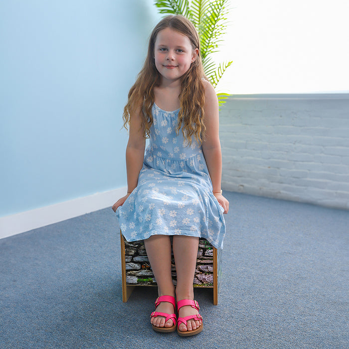 Young girl in a blue floral dress sitting on a small stool against a light blue wall.