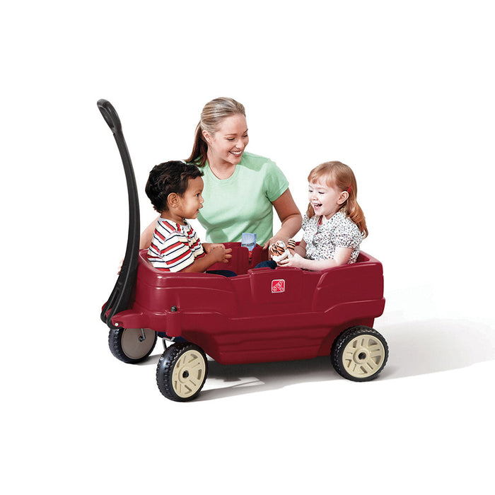 Two children, a girl and a boy, sitting in a red two-seater plastic wagon with a black handle with an adult supervising